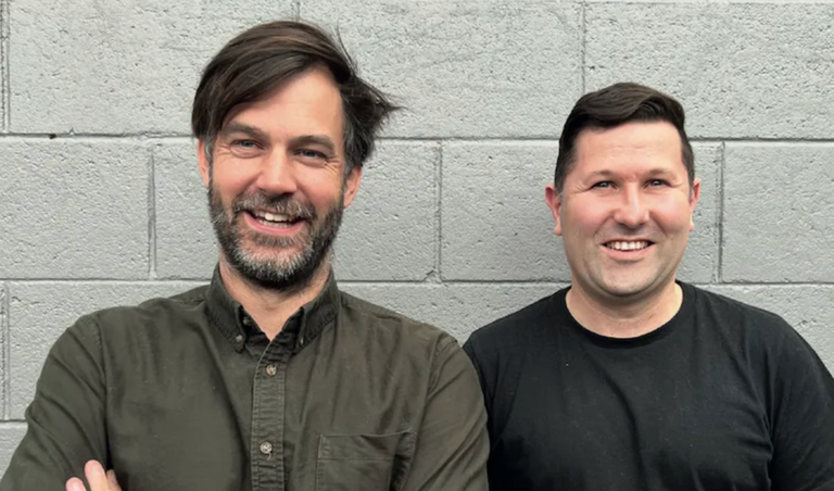 Five Four Entertainment founders Craig Lock and Ross Osman smiling together in front of a grey concrete brick wall for a promo portrait.